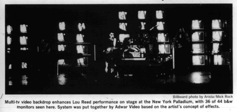 A black and white photo showing Lou Reed on stage in front of flickering TVs