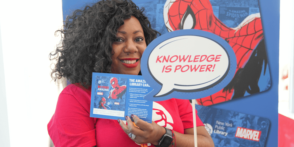 Smiling librarian in a red t-shirt holds up a Spider-Man library card and a comic book–style speech bubble that reads Knowledge Is Power!