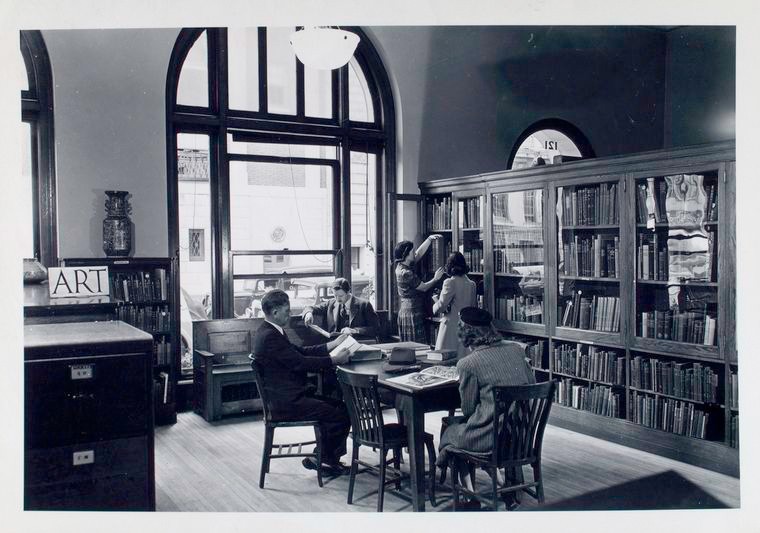 Librarian at bookshelf with girl, others reading