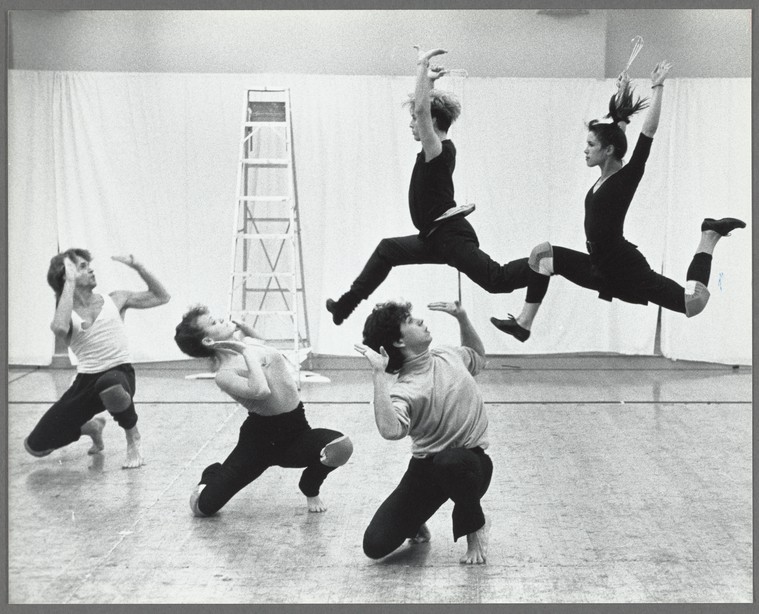 two dancers leap in the air while three more are crouched on the ground in a rehearsal space