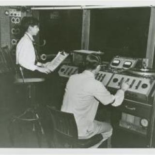 Black and white photo of two people at a recording booth.