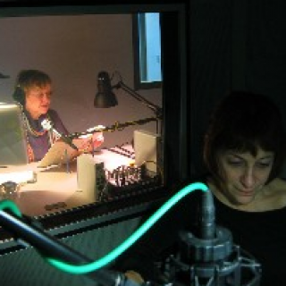 Photo of two people in a recording studio: A person sits in a darkened foreground near a microphone and in the background, another person sits underneath a light and reads from a piece of paper into a microphone