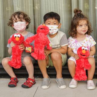 Photograph of three diverse children in masks, sitting in front of a window, and holding Elmo dolls