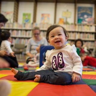 Photo of a small toddler in the foreground wearing a frilly, white, long-sleeved tee sitting and smiling on a colorful checkerboard mat amongst other caregivers and children blurred in the background