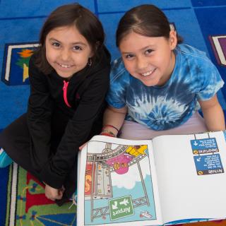 Photo of two young girls sitting on a colorful blue carpet, smiling at the camera, and holding an open picture book