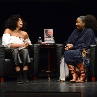 : Activists Sasha Bonet and Tarana Burke are seated in chairs across from each other on stage at the Schomburg Center. There is a small round table between the two women with a copy of Burke’s book, Unbound: My Story of Liberation and the Birth of the Me Too Movement.