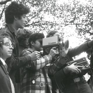 A black and white photo of several kids videotaping something off the screen, they stand in front of a tree canopy