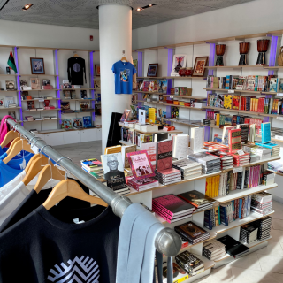 Interior view of the Schomburg Shop. There are books on short and tall shelves. In the lower left corner, a partial view of clothing rank featuring items with Center's logo.