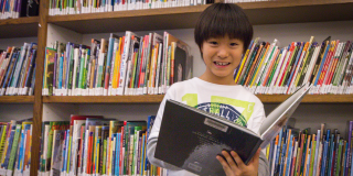 Young boy smiles and holds a book in a room full of bookshelves.