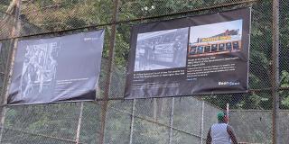 Two brown banners with two black and white photos and one color photo hanging on a fence in a park with a man in the background