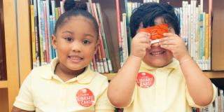 Two children in yellow polo shirts stand in front of a bookshelf; one child holds up a red NYPL card and they are both wearing stickers that read: I got a Library Card! 