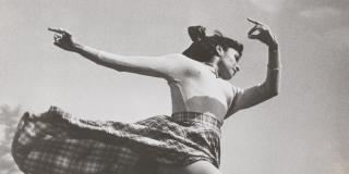 Black and white photo of a dancer in a long sleeve shirt and long plaid skirt leaping in field.