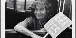 Photograph of old woman through an open window. She is smiling and holding a sign that reads "ERA YES"