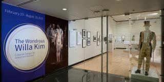 Entrance of the exhibition with a purple title graphic to the left and a mannequin with a decorated suit and top hat in the window to the right.