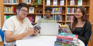 Three teens using a laptop and reading at a table in front of a wall of bookshelves.