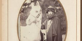 Sepia-toned image of man in robes standing next to a white horse