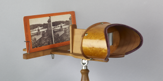 Photo of a stereoscope, which features a wooden handle and a viewing window for users to look through in order to see a card with two nearly identical images of Bethesda Fountain in Central Park.