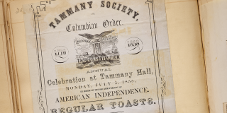 Photo of a historic scrapbook featuring records from Tammany Hall; the yellowing, stained page features text that reads: Annual Celebration at Tammany Hall. 