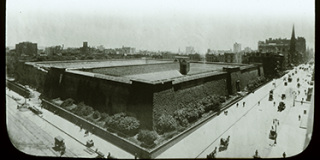 Black-and-white lantern slide of the Croton Reservoir with hedges on around the sides of the reservoir and a view of Fifth Avenue filled with carriages and pedestrians.