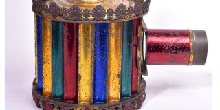 a round hatbox-sized container has two tubes coming from it: one going up and one pointing out through the side. The lantern itself is brightly striped with red, blue, green, and yellow