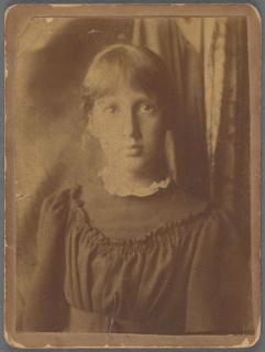 A sepia portrait of a teenage girl with bangs and hair pulled back, wearing a high-collared top and posing in front of a curtain.