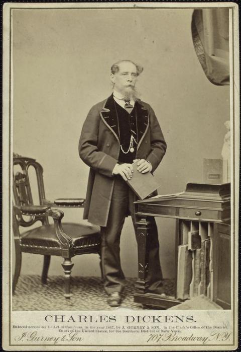 Charles Dickens is pictured standing at a writing desk and holding a book. He looks just off center and a watch hangs from a chain on the vest he wears inside a large lapelled coat. 