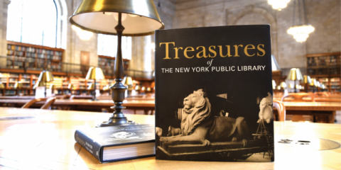 Photo of an upright book called Treasures of The New York Public Library, propped up next to a gold lamp on a table inside of the Rose Main Reading Room.