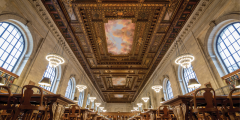 Photo of the Rose Main Reading Room from a low perspective that shows an ornate ceiling, large arched windows, chandeliers, and rows of wooden tables. 