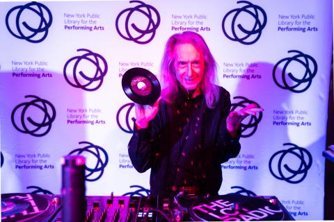 Purple and blue lights shine on a man with long gray hair as he holds up a 78 vinyl record before a set of turntables.