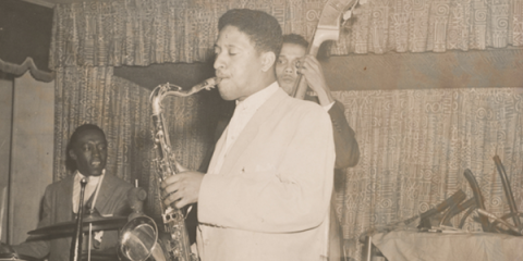 Sonny Rollins is standing in the foreground of the photo playing the saxophone. In the background is drummer Art Blakey and double bass player Tommy Potter. 