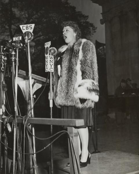 A black and white photo of a woman outside singing into several press microphones.